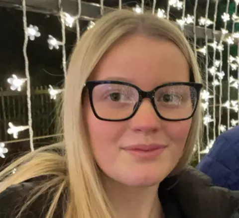 Grace Darnbrough Grace Darnbrough smiles at the camera with fairy lights behind her. She has straight blonde hair and wears black glasses and a black puffer jacket.