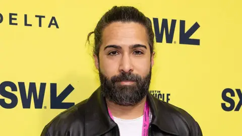 Getty Images Savan, a man, wearing a black leather jacket, with the background a yellow boarding with the letters SW on it.