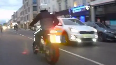 Met Police Motorcyclist riding bike in CCTV image taken from behind. He is wearing a white helmet and dark clothing with a white reflective line going down his trousers.