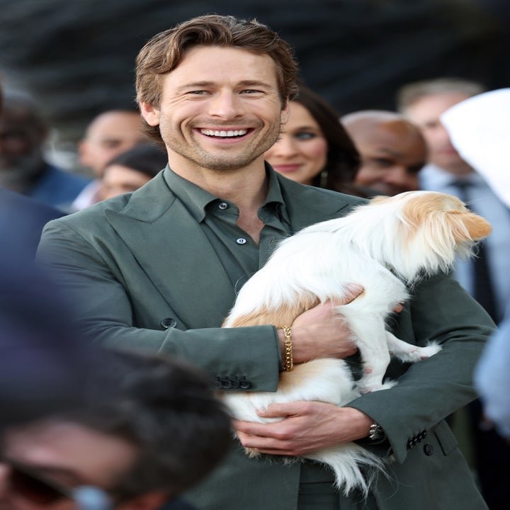 Glen Powell is smiling and holding a small dog at an event. He is wearing a suit