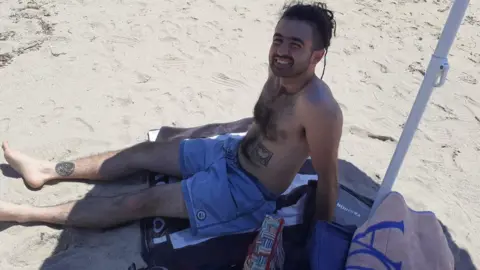 Handout A man lying on a sandy beach wearing shorts, smiling up at the camera