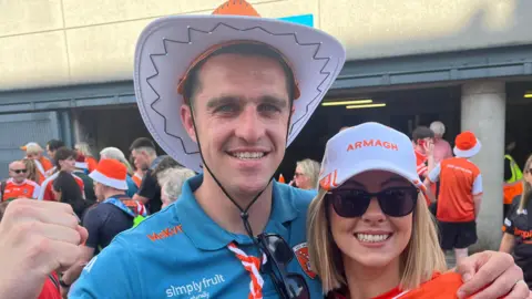 A man is wearing a cowboy hat and a blue Armagh top. His arm is around a woman wearing a cap, sunglasses. and an orange jersey.