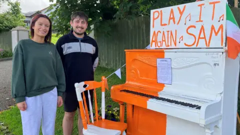 Chloe and Ogie Marks standing in front of a piano, coloured in orange and white