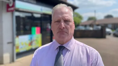 Laura Foster/BBC Carl Foster in a shirt and tie stood outside the shop where Carol Morgan was murdered