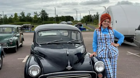 Steph Holloway Steph Holloway leans against a black classic car, with her hand resting on the bonnet. She has red hair styled in a beehive and is a wearing a checked two-piece suit with a blue shirt