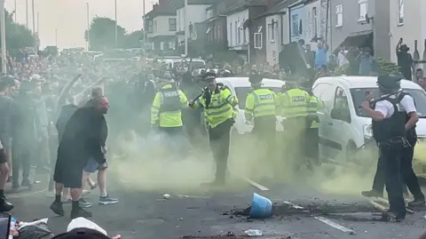 Richard McCarthy/PA Media Hundreds of protesters gather in a street and are faced by a small number of police officers in hi-vis jackets and uniform holding batons