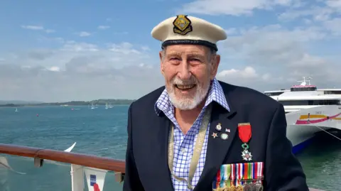 PA Media George Winter from shoulders up wearing a suit adorned in medals and wearing a sailors hat. He is stood on a boat, with other boats in the background as well as the water and shoreline in the distance.