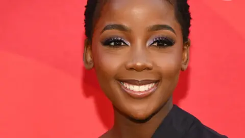 MICHAEL TRAN/AFP A young, smiling woman with cropped hair and a black outfit stands in front of a red background posing for photos.