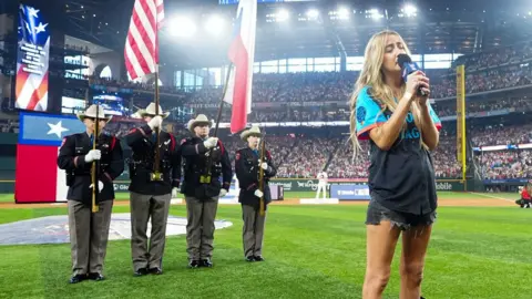 Getty Images Ingrid Andress singing on the baseball field with the crowd and servicemen holding flags in the background