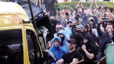 Getty Images A crowd of people, some with their faces covered, standing in front of a police van. Some appear to be throwing a wheelie bin while many others are holding up their phones to film the scene