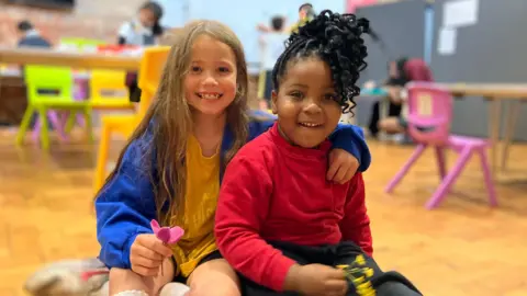 BBC A child with long, light brown hair, and a yellow and blue top, has her arm around another child with black curly hair. They're both smiling and sitting on a wooden floor inside a holiday club.