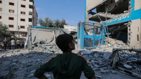 EPA A Palestinian boy stands near the rubble of a damaged Unrwa-run school in Nuseirat refugee camp, central Gaza, following an Israeli air strike (14 July 2024)