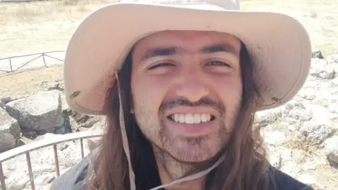 Handout A bearded man with a cream hat on, with long brown hair stood infront of rocks