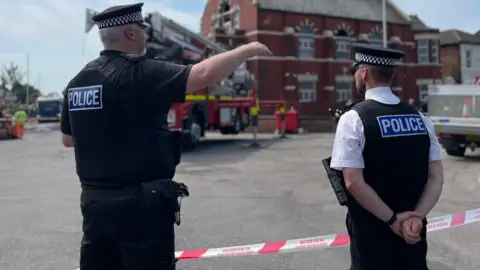 Merseyside Police Two police officers, one in a black uniform and one wearing a black stab vest and a white shirt, stand in front of a fire engine on a street in Southport