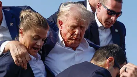 Getty Images Bleeding from the ear, Donald Trump is helped off stage by male and female secret service agents.