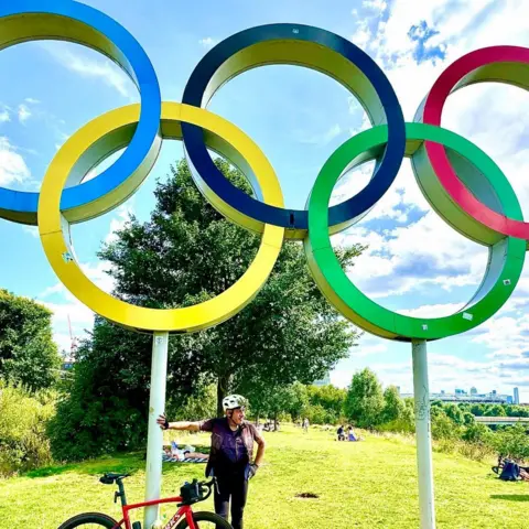 Nico Georgiou Mr Georgiou with his bike standing in front of an Olympic rings monument