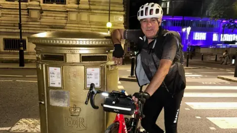 Nico Georgiou Nico Georgiou standing next to a gold letter box with his bike