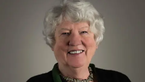 Head and shoulders shot of a smiling Dame Elan who has white hair and is wearing a dark top