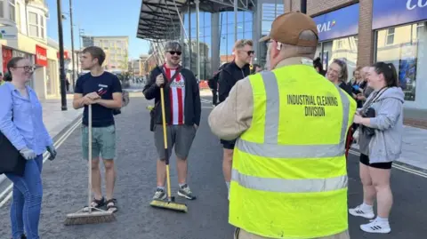 A group of people with brushes preparing to clean up the streets after disorder. A man in a yellow jacket with the logo Industrial Cleaning Division is standing in front of them.