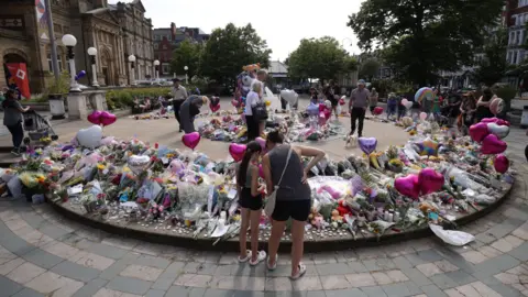 PA Media People stand and look at flowers, balloons, cuddly toys and other tributes left outside the Atkinson art centre in Southport