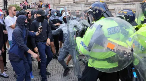 Reuters a group of police officers in riot gear and carrying clear shields push forward into a group of men in casual clothing, most of whom have their faces covered