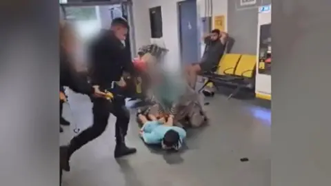 Handout A man lies on his front on the ground with his arms by his sides while an officer in uniform holding a taser draws his leg back