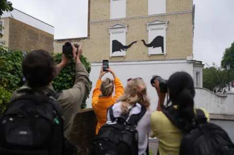 Yui Mok/PA Media People photograph a new artwork depicting two elephants poking their heads out of blocked-out windows unveiled by Banksy 