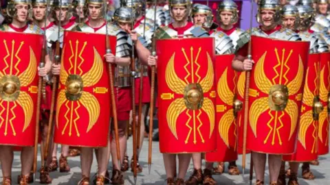 Getty Images About 15 men dressed as ancient Romans in armour and carrying spears and red and yellow shields