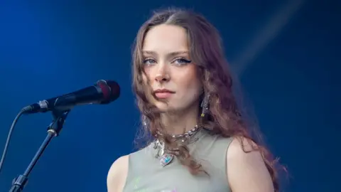 Getty Images Holly Humberstone on stage. Holly's a 24-year-old woman with long curly brown hair worn loose. She has blue eyes, rimmed with a thick black eyeliner. She wears a high-necked sleeveless grey top with a chunky silver necklace and is pictured against a blue backdrop.