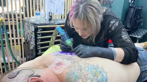 BBC/JESS LORD Image of Naomi Dalby tattooing the back of deaf client, Paul Gardner.