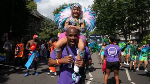 PA Media A girl in a feathered costume is carried through the street on her dad's shoulders