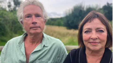 Max and Liz Waldron. They are sitting on a bench surrounded by fields and trees. Max is wearing a green gingham shirt while Liz is wearing a black shirt.