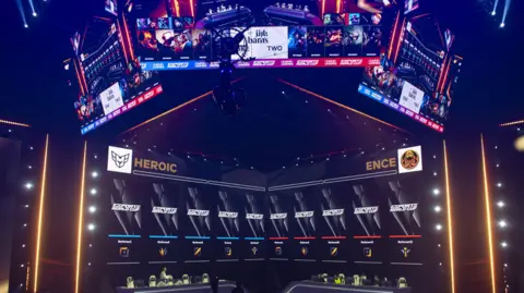 Getty Images Stands are prepared for visitors during the opening ceremony of 2024 Esports World Cup in Riyadh, Saudi Arabia. The stage is lit purple with two long desks side by side in front of a score board. 