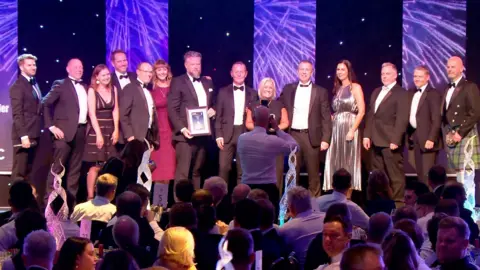 Housebuilder More than a dozen people on stage posing for a picture as they accept an award. The dress code is black tie. A phographer in a white shirt is standing in front of the stage with his camera raised and other guests are seated at tables in the foreground.