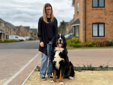 Megan St Denis standing outside and looking down at her dog Teddy, who is sitting. The blurred background shows a residential street with vehicles parked on driveways.