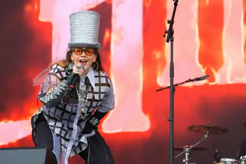 Getty Images Pop artist CHINCHILLA performs wearing a silver top hat, a checked shirt and orange tinted sunglasses. She also wears black trousers whilst holding a microphone, singing on stage.