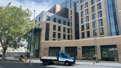 A brand new building with a tree outside, and a van parked in front of it. The walls of the building are light brown and some of the facia is a dark grey. There are also multiple glass windows visible