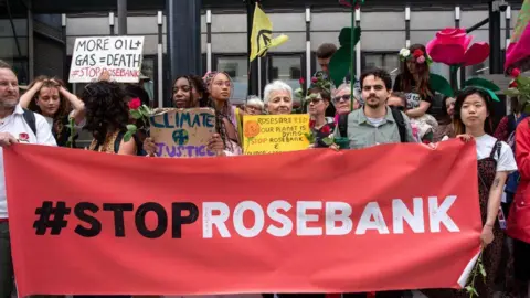 PA Media Group of protestors holding small signs standing behind a large red stop rosebank banner.