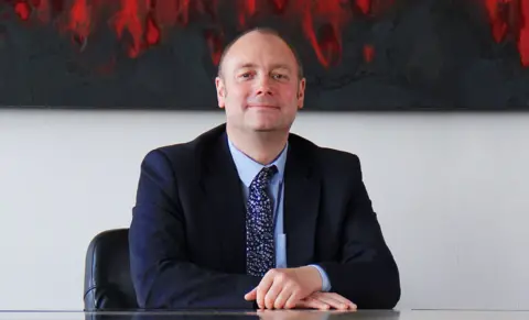 Hartlepool College of Further Education Darren sits at a desk with his hands folded on top of one another. He smiles at the camera and is wearing a dark blue suit with a floral navy tie.