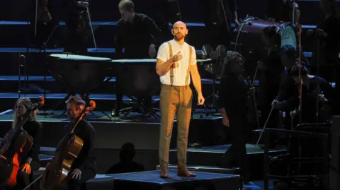 BBC/Sisi Burn Thomas Simper on stage at the Royal Albert Hall, surrounded by the Aurora Orchestra.