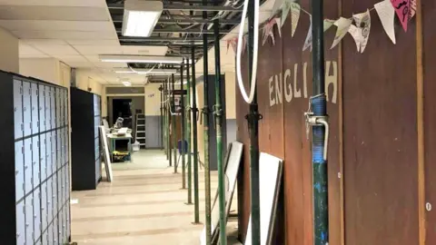 Altrincham College A school corridor with 'English' written on one wall and lockers lining the other. Ceiling panels have been removed and metal poles prop up the ceiling.