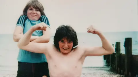 Courtesy of Patrick Jones Teenager Nicky shrugs his shoulders behind his older brother Patrick who flexes his muscles while on a family holiday on a beach in the UK   