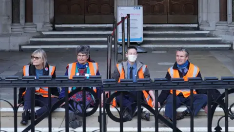 Ian West/PA Media (Left-right) Theresa Norton, Dr Diana Warner, El Litten, and Steve Pritchard, sitting outside the Royal Courts of Justice in London. They are members of Insulate Britain and they glued themselves to the ground outside the court.