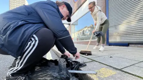 BBC Man in track suit and cap crouching down and woman on pavement with a brush sweeping glass into a bin liner.