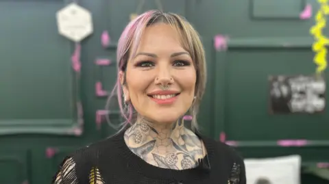 BBC/JESS LORD Naomi Dalby has pink and blonde hair, a septum and nose piercings, and tattoos on her neck. She is smiling at the camera and wearing a black top.