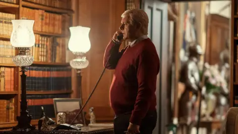 Amazon MGM Studios Michael Sheen, playing Prince Andrew, in a red jumper stands next to a desk, holding a telephone to his ear.