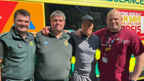 West Midlands Ambulance Service Four men stand smiling in front of an ambulance, two of them wear paramedics' uniforms and one wears scrubs