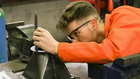 Hartlepool College of Further Education A student wearing a bright orange jacket and protective goggles works on a piece of machinery.