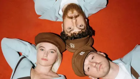 Elliot Morgan Promotional photo for Clean Bandit, showing the band lying on a red carpet in a clockwise arrangement