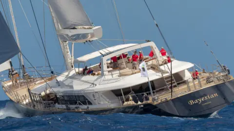  Perini Navi Press Office The Bayesian yacht seen sailing at sea, the large yacht appears to be turning in the water with a number of crew members visible on deck wearing red shirts. The name of the boat is visible on the rear on the boat 